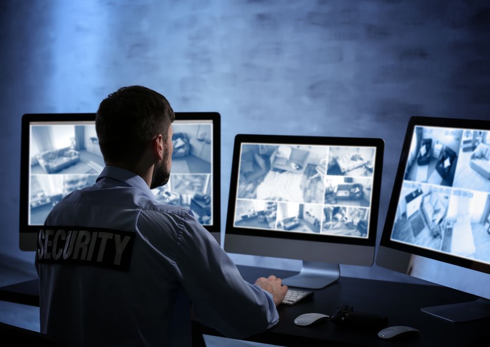 A security guard is sitting in front of three computer monitors.