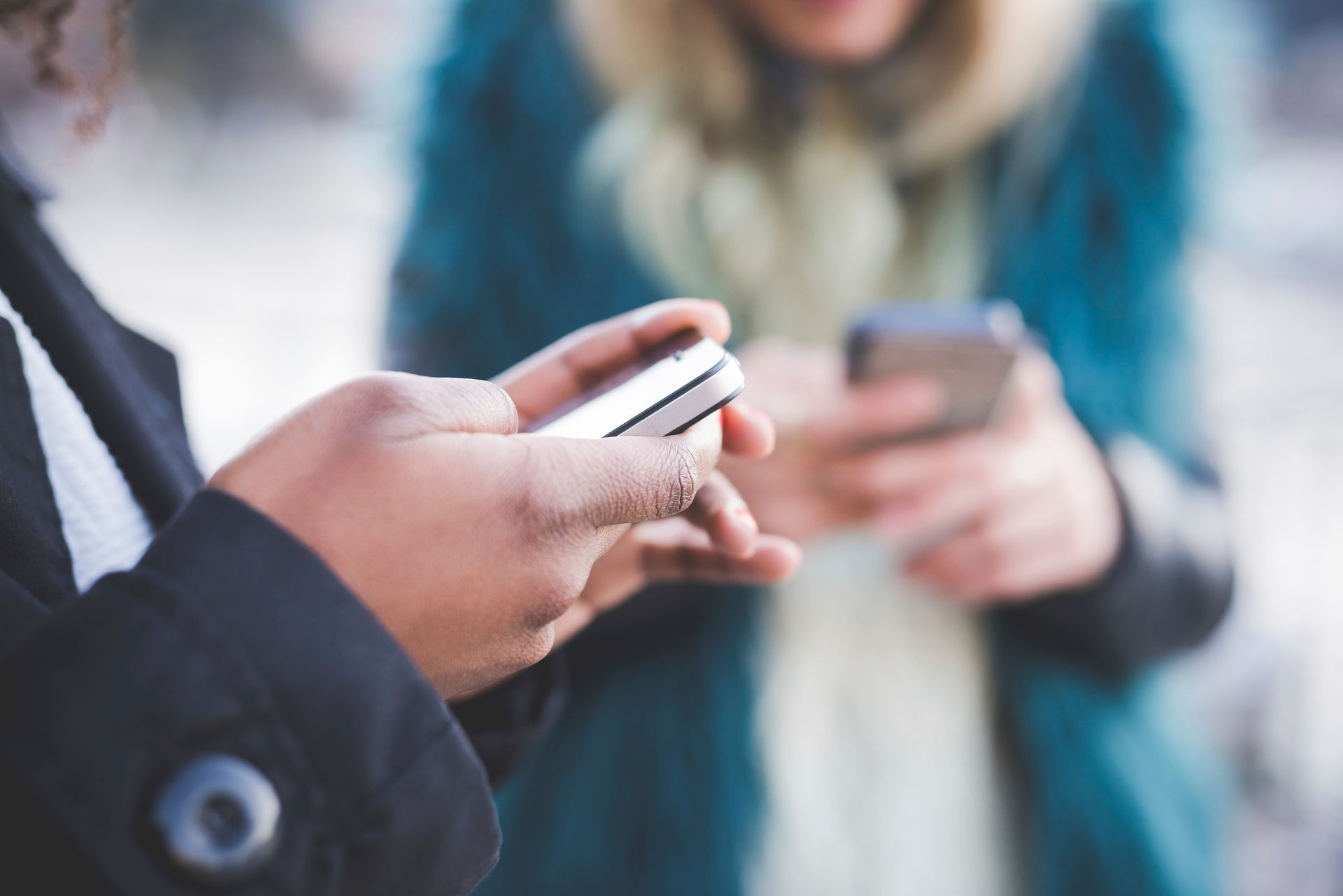 Two people using smartphones outdoors. Focus on hands holding phones; one in black coat, other in teal coat.