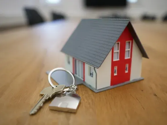 Model house with keys on a wooden surface, symbolizing homeownership. Red and white facade, gray roof.