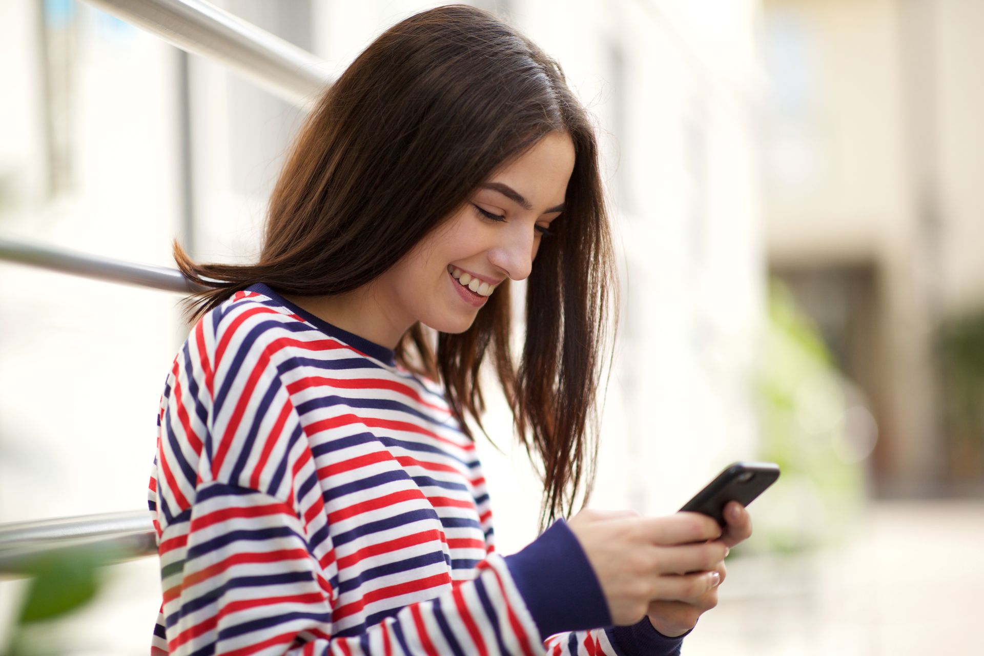 Woman smiles while looking at a phone. She wears a striped shirt and leans against a railing.