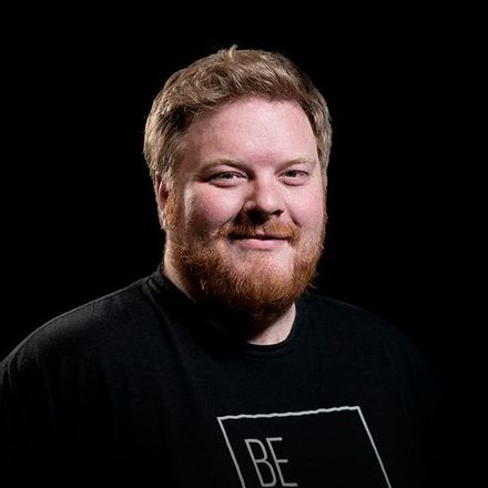 A smiling man with red beard and hair against a black background. He wears a black shirt with a white graphic.