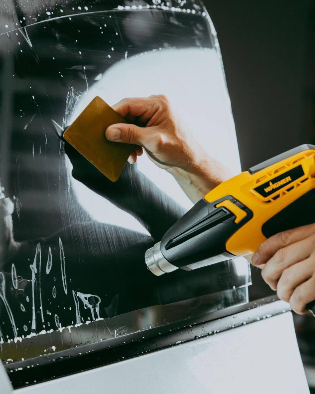 Person applying window tint with a scraper and heat gun.