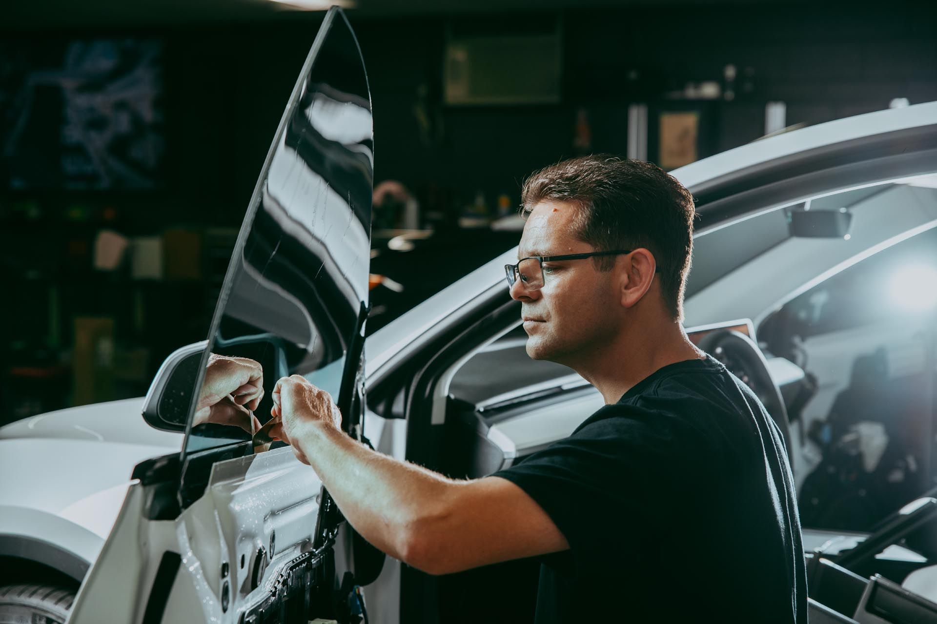 Man working on a car door in a garage. He is wearing glasses and a black shirt.
