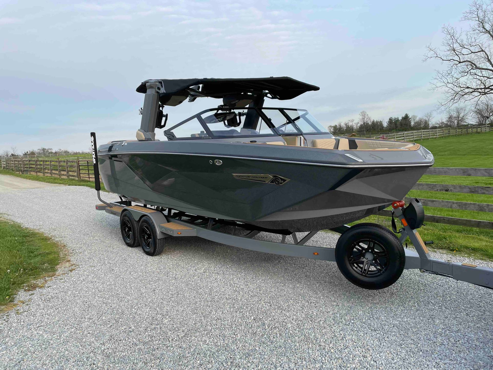 Gray wake boat on a trailer, parked on gravel. Features a black tower and a light brown interior.