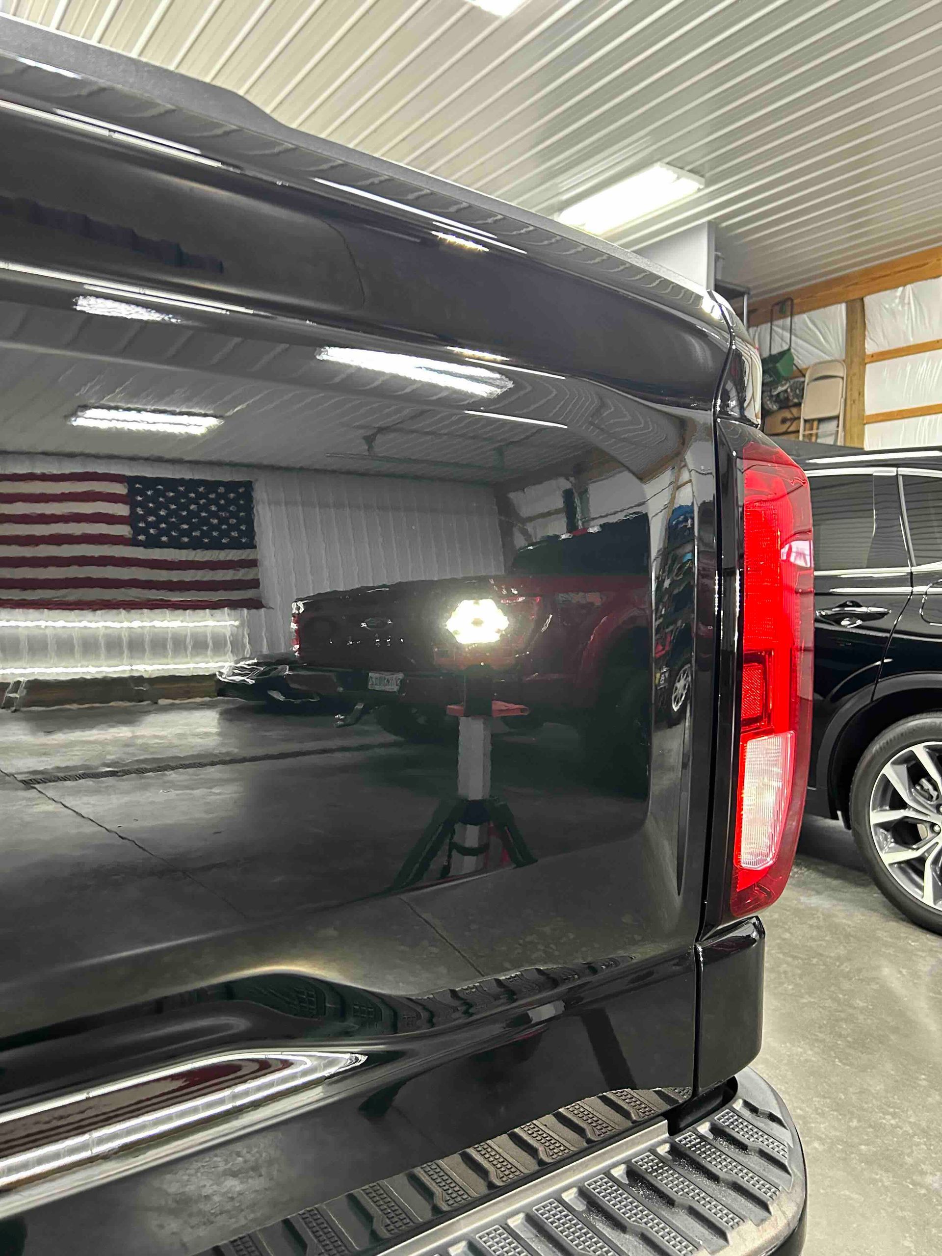 Black truck bed reflecting a red vehicle and American flag in a well-lit garage.