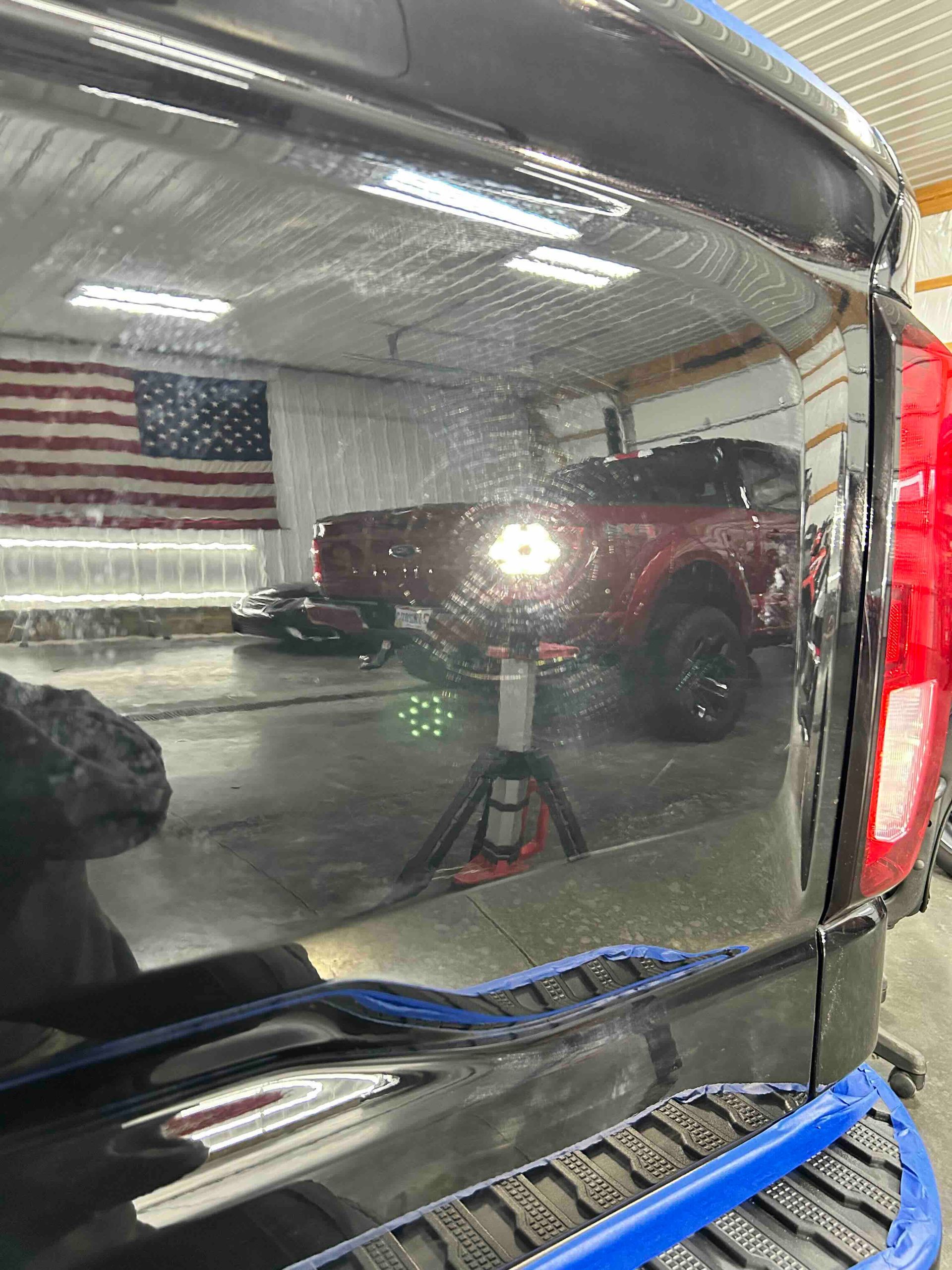 Black vehicle's glossy paint reflecting a shop interior with a red SUV, an American flag, and a work light.