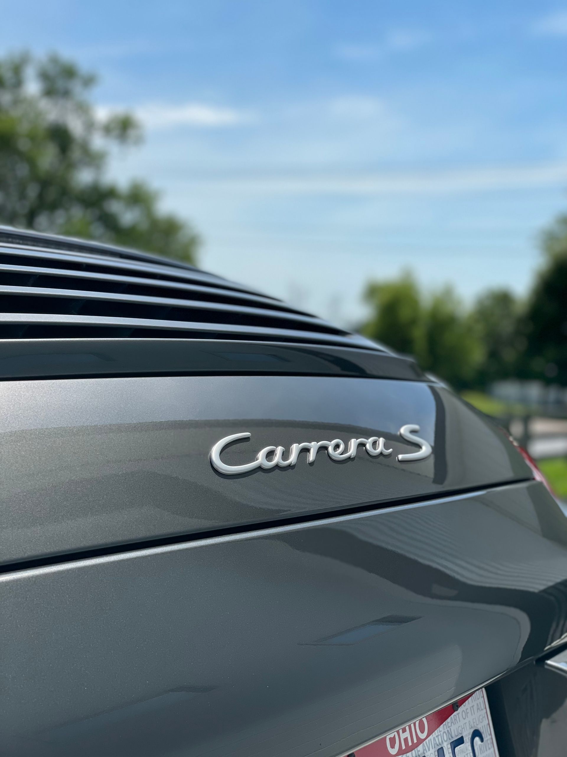 Close-up of a gray Porsche Carrera S badge on the car's rear, with a blurred background of trees and sky.