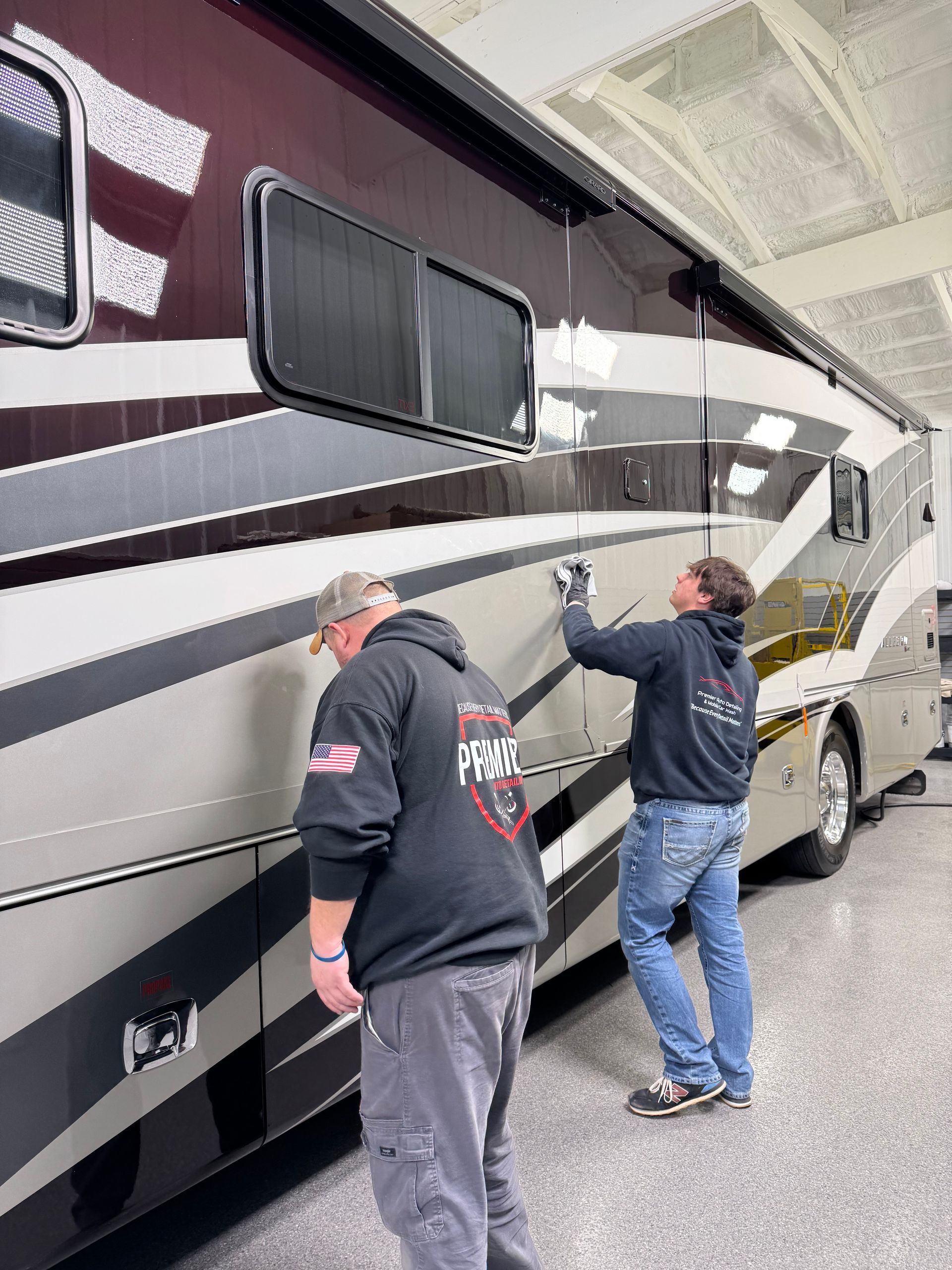 Two men working on the side of a large RV; one cleans windows, the other examines.