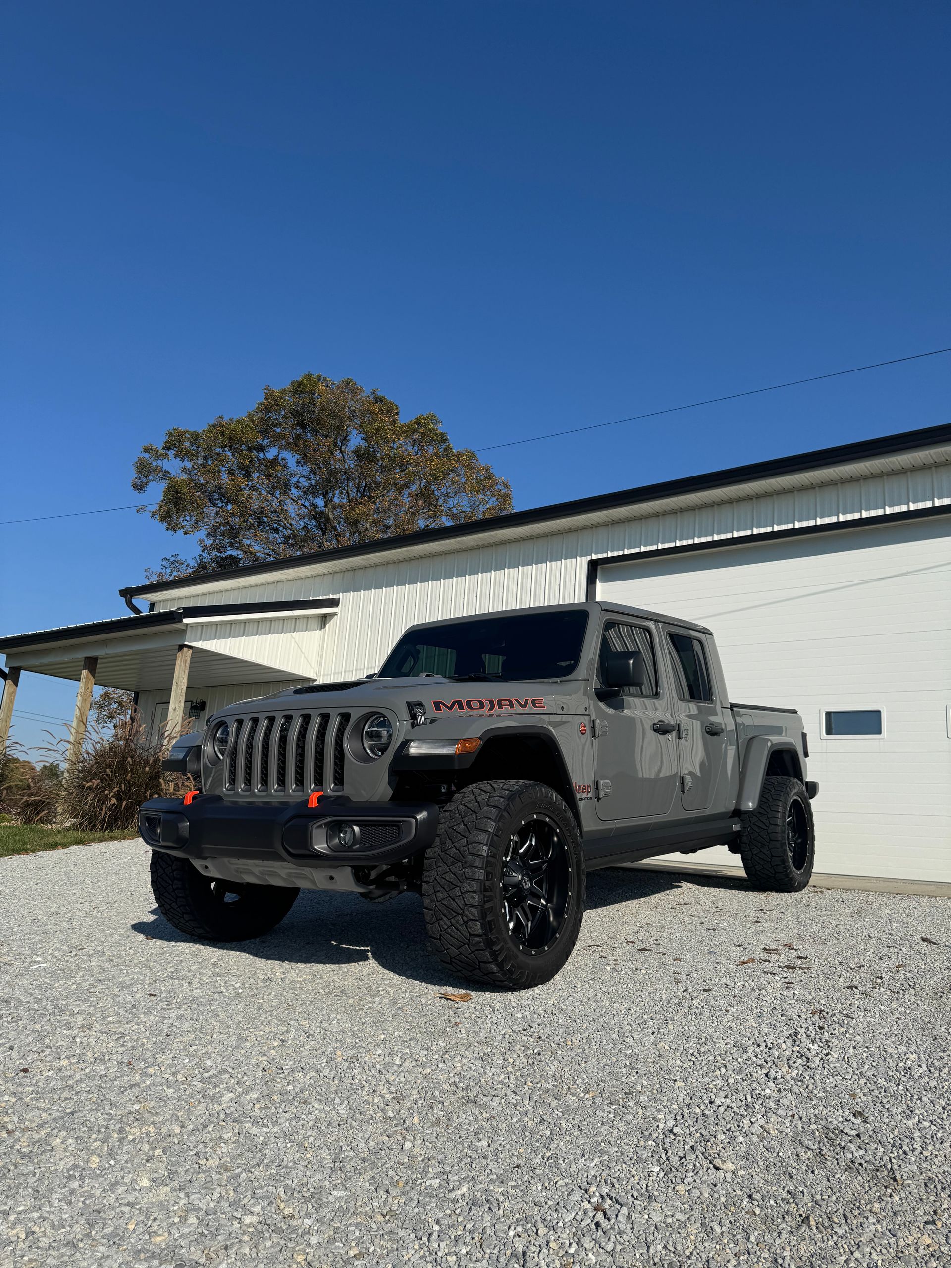 Gray Jeep Gladiator parked in front of a white building on a gravel driveway, under a blue sky.