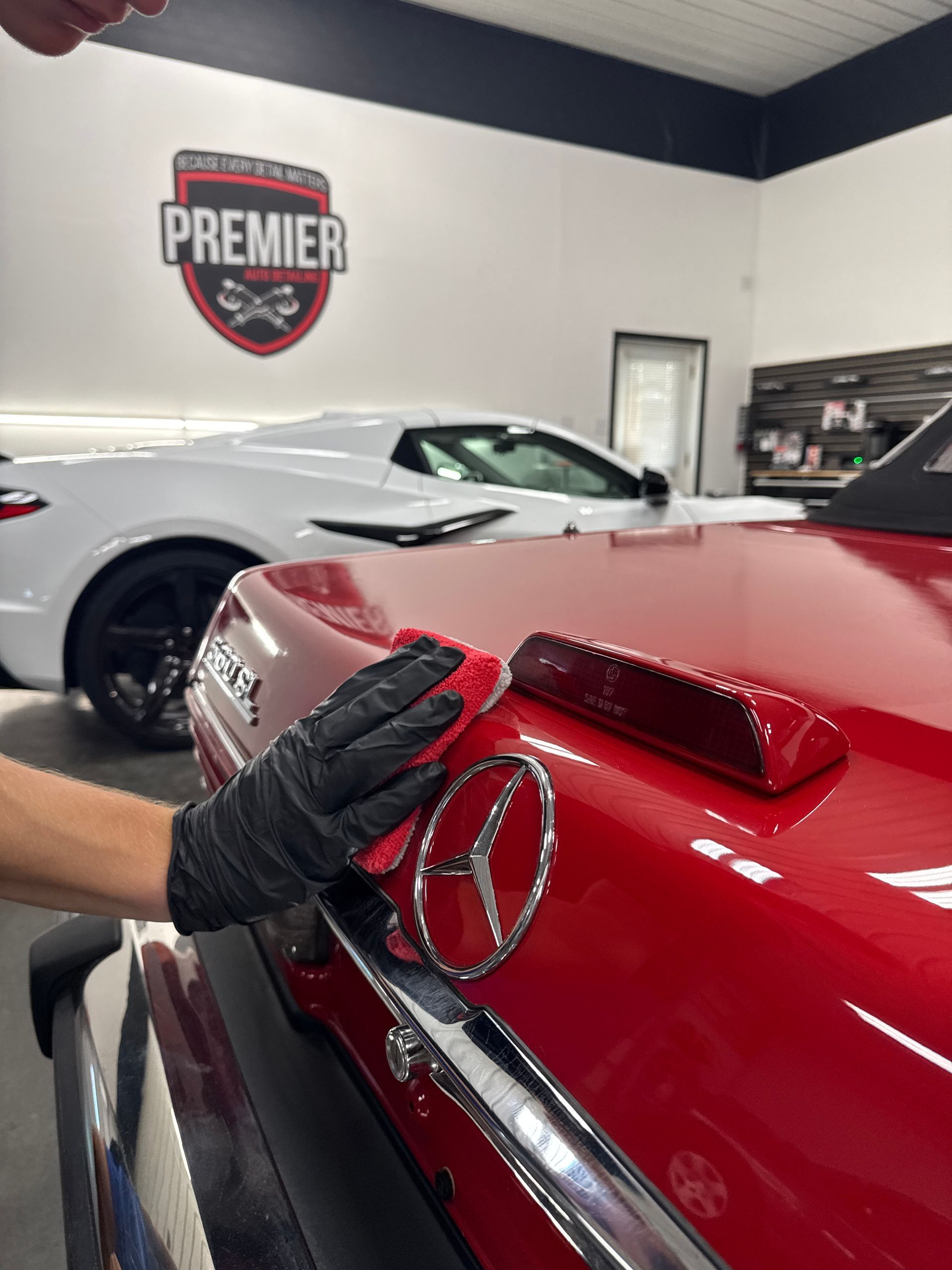 A gloved hand polishes a red car's trunk with the Mercedes emblem, a white car in the background.