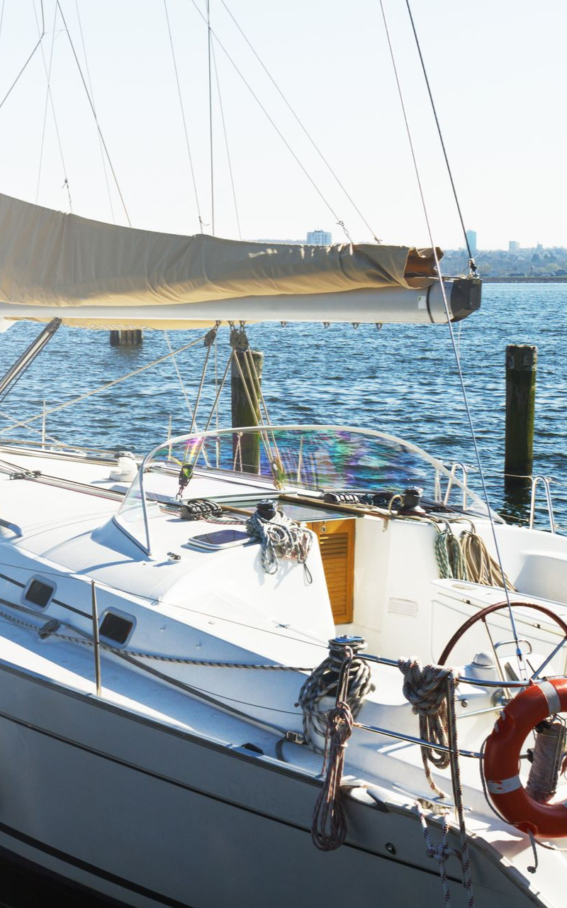 Sailboat docked by wooden posts on water; beige sail, white hull, life preserver visible.