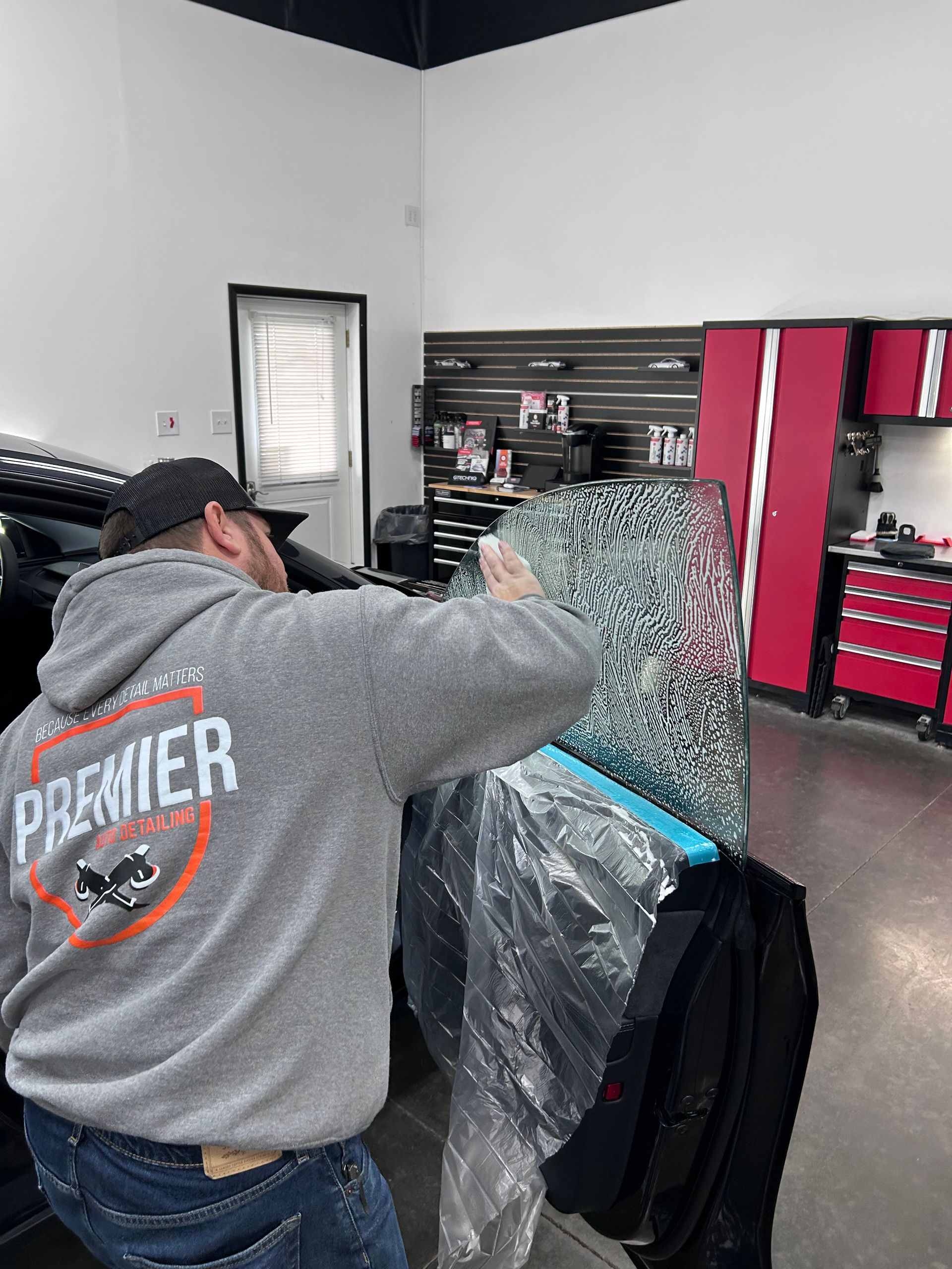 Man applying tint to a car window in a shop. He wears a grey hoodie with a logo and is hunched over his work.