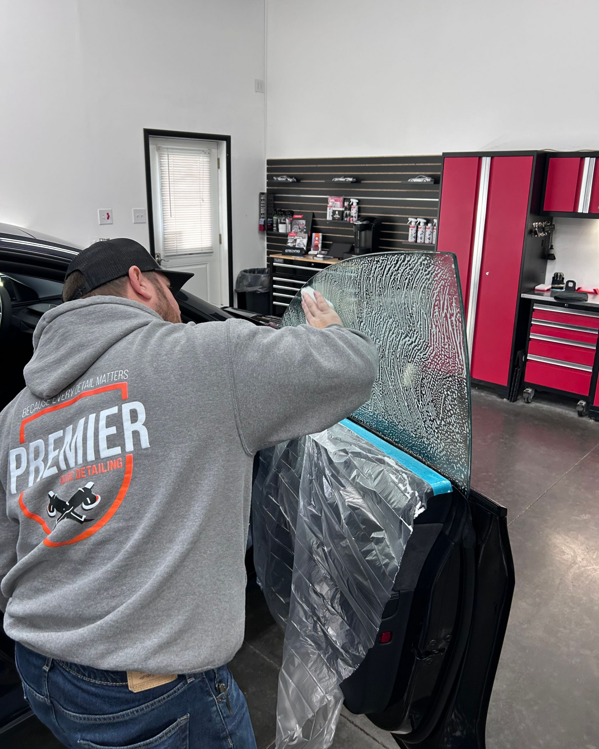 Person applying window tint to a car window, inside a shop. Gray sweatshirt, red tool cabinets in background.