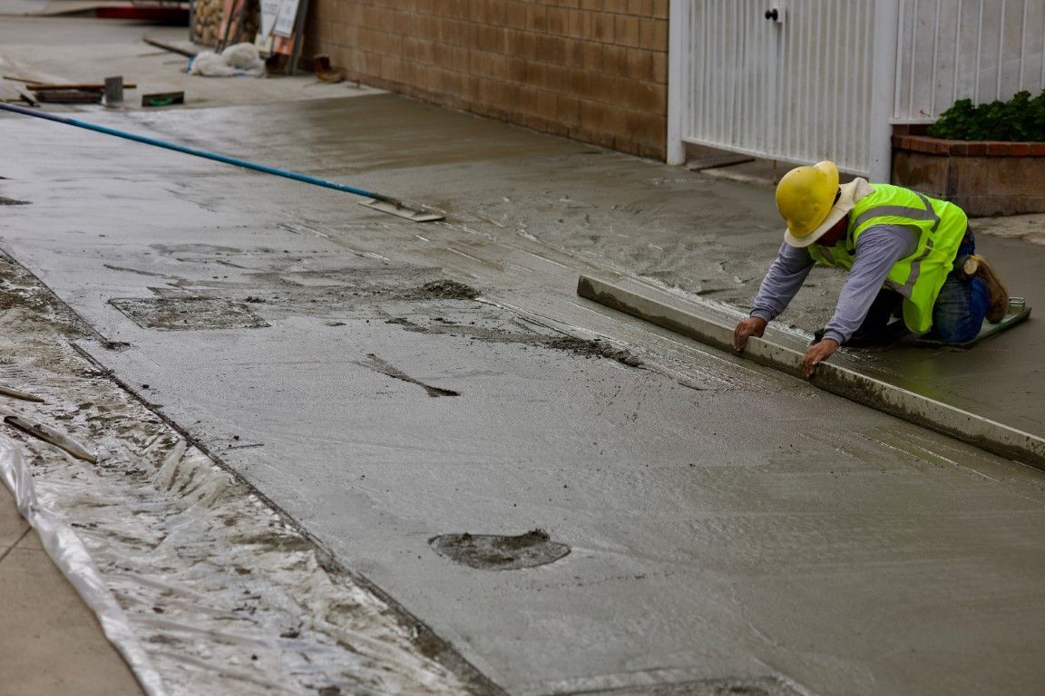 A construction worker in a yellow vest and hard hat smoothes wet concrete with a tool. 