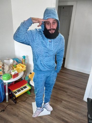A man with a beard wearing a blue hoodie and sweatpants is standing in a living room.