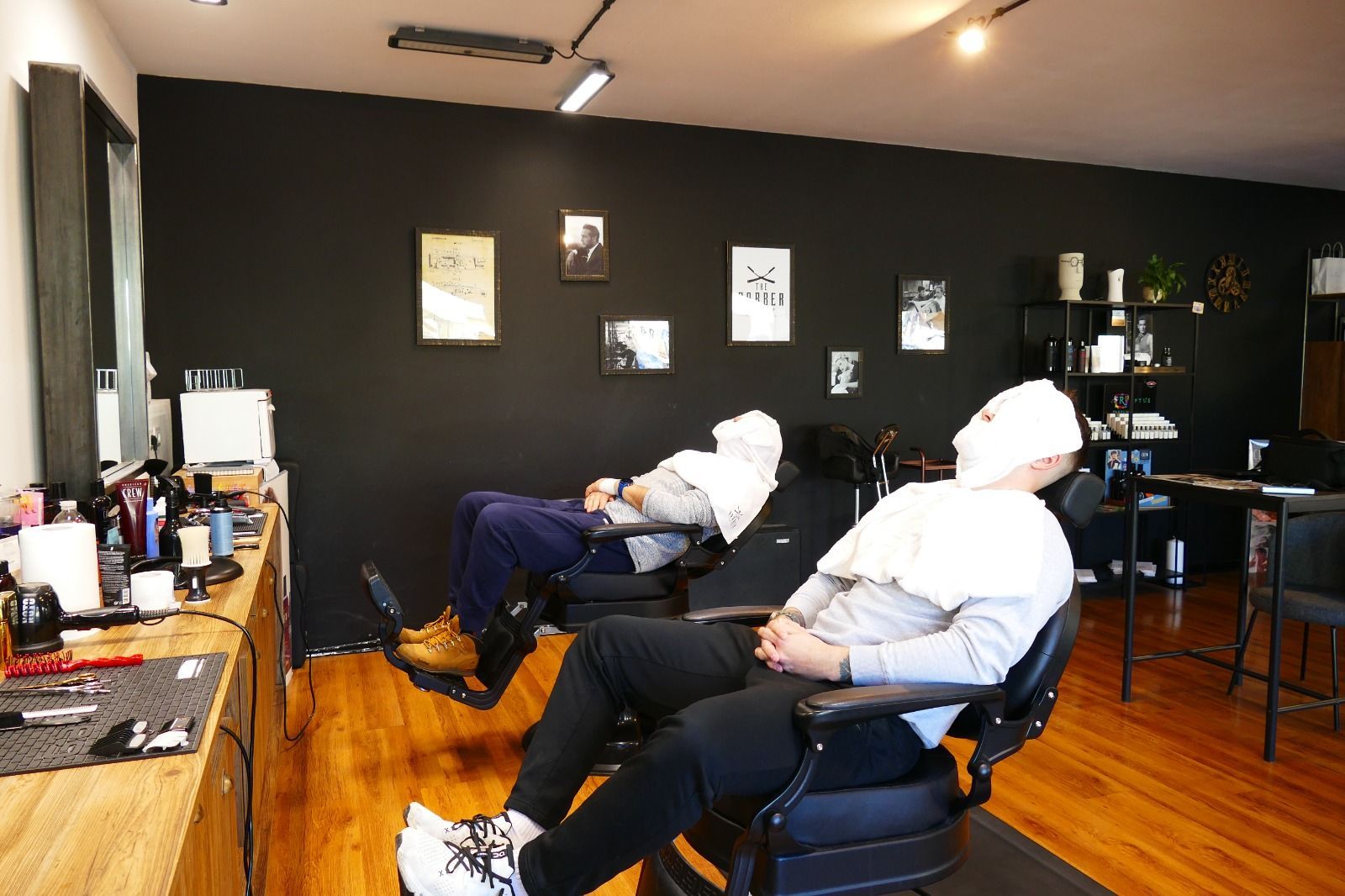 Due persone sdraiate su poltrone da barbiere, coperte da asciugamani. Interno di un barbiere con pareti nere.