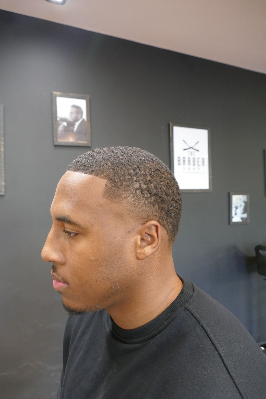 Uomo con taglio di capelli sbiadito visto di profilo in un barbiere, con un'opera d'arte incorniciata su una parete scura.