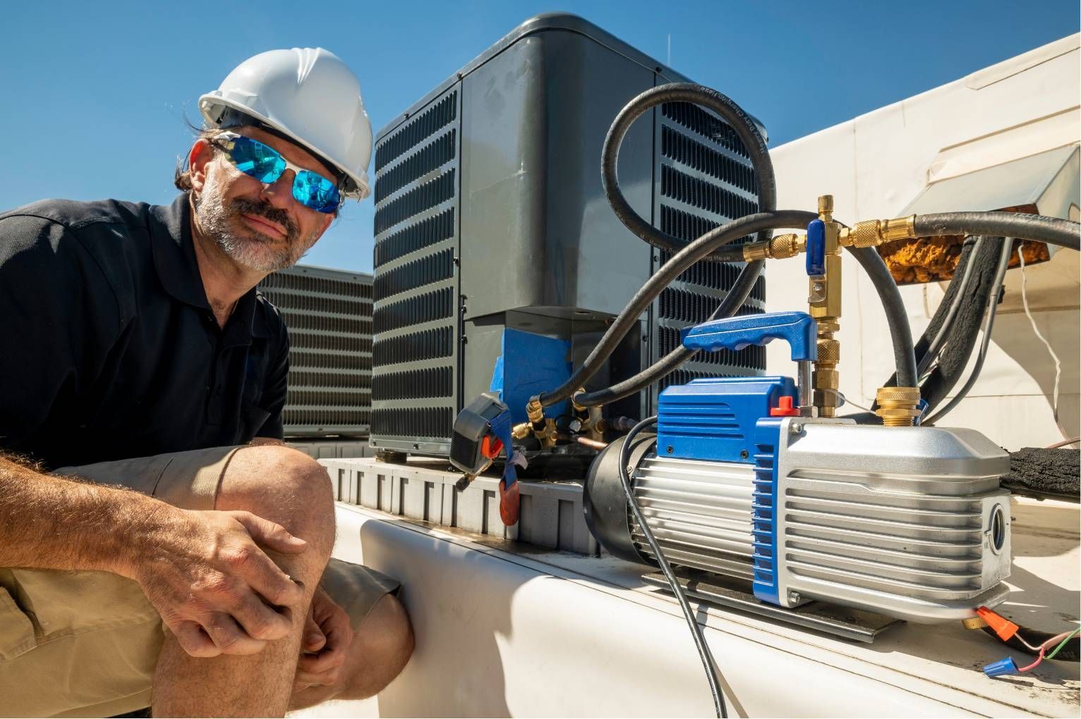 Worker in hard hat inspecting rooftop HVAC equipment beside a large building.