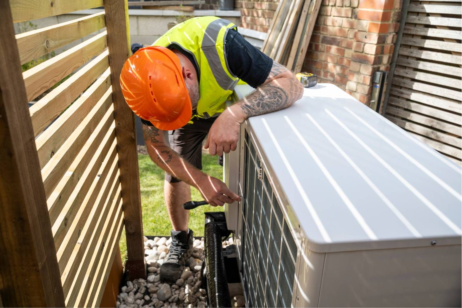Worker in orange hard hat installing equipment beside a white outdoor unit in a narrow alley.