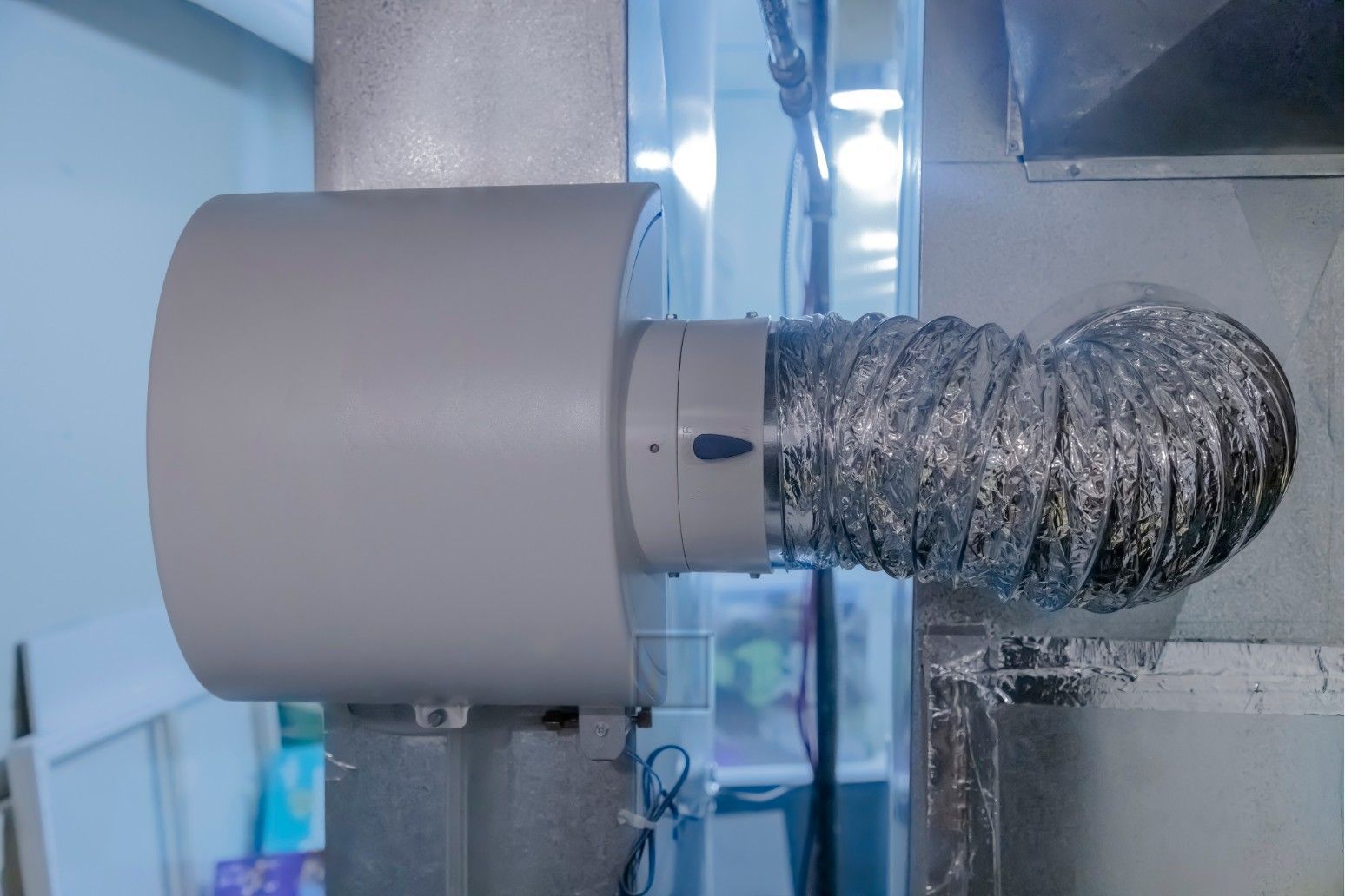 White ventilation fan connected to a silver flexible duct in a room near a window