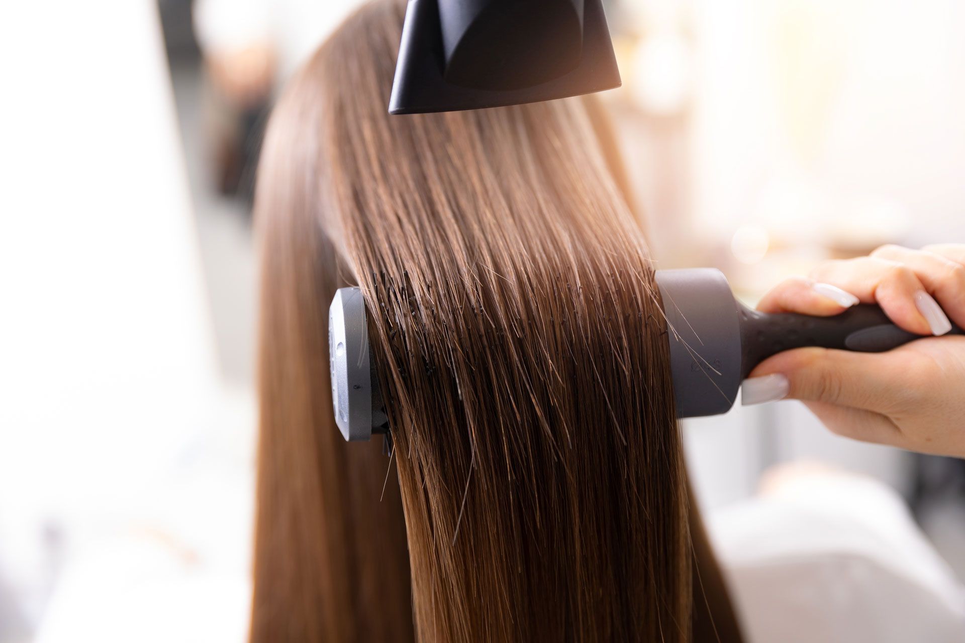 Blow dryer styling long, straight, brown hair with a round brush in a salon setting.