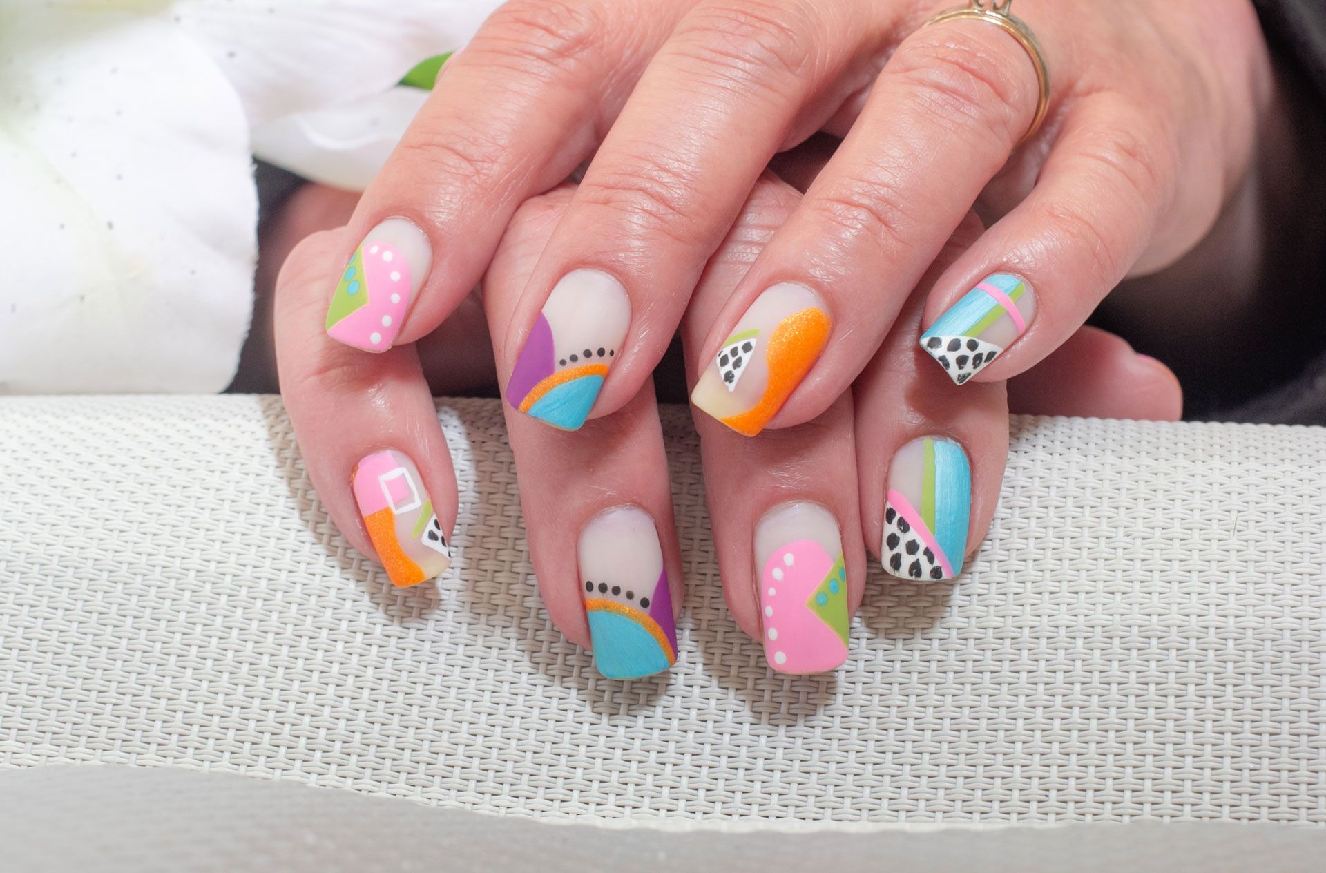 Hands with colorful abstract nail art: pink, orange, blue, green, and black designs on a neutral base.