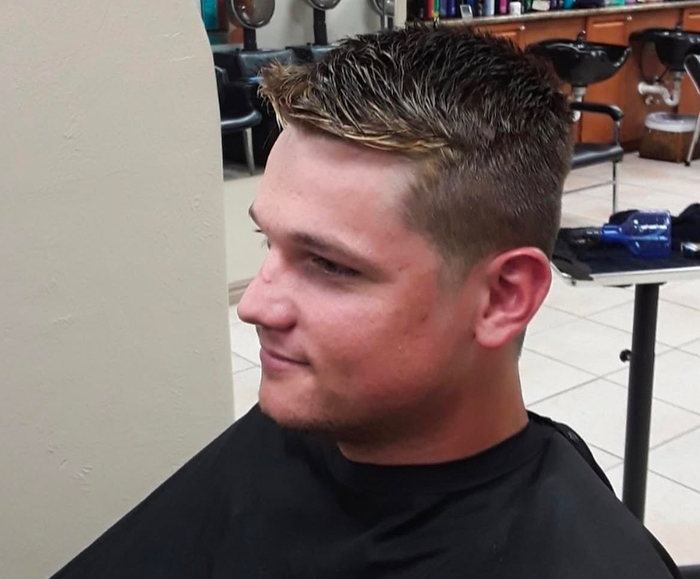 A person in a salon cape with a short, textured, dark-brown haircut, viewed from the side profile against a salon backdrop.