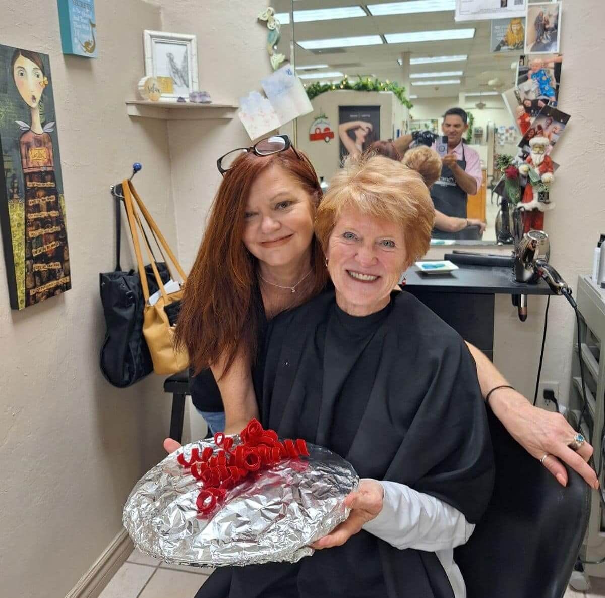 Two women smiling in a salon, holding a wrapped tray.
