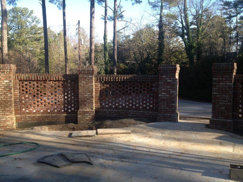 A brick wall surrounds a driveway with trees in the background