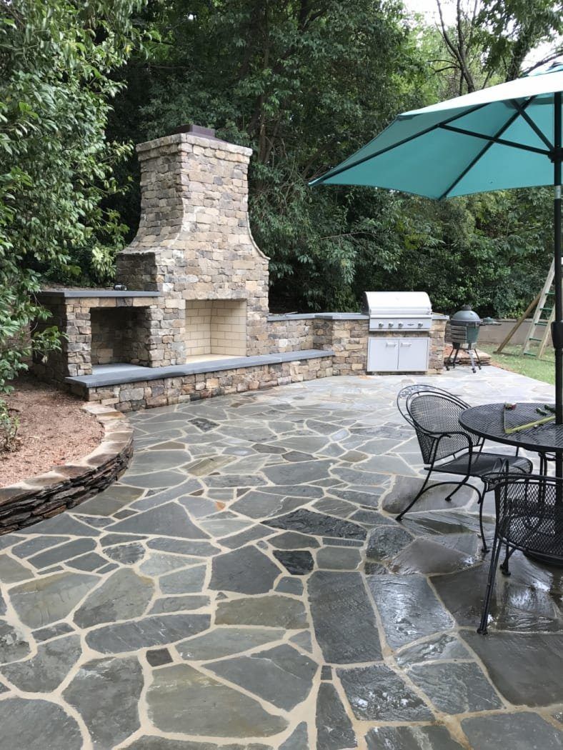 A stone patio with a fireplace, grill and umbrella