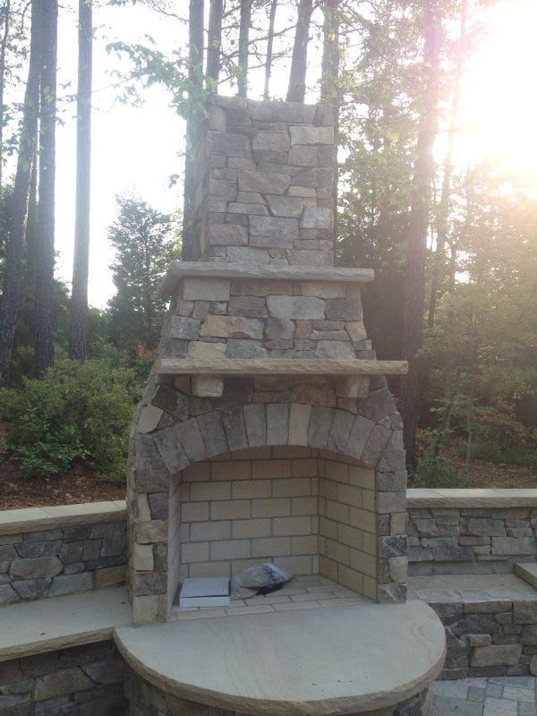 A stone fireplace in the middle of a forest with the sun shining through the trees