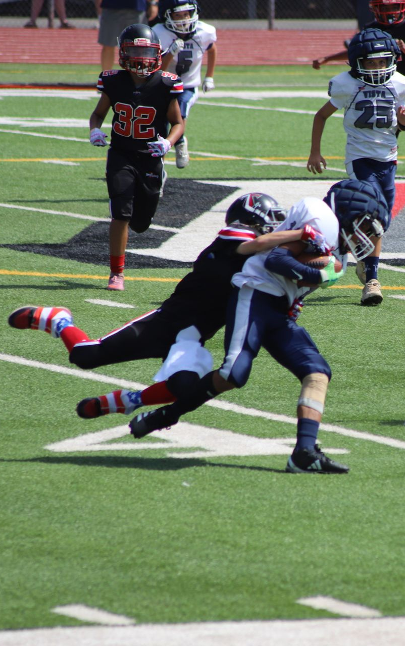 Football players in action on a grassy field; one runs with the ball, pursued by two opponents.
