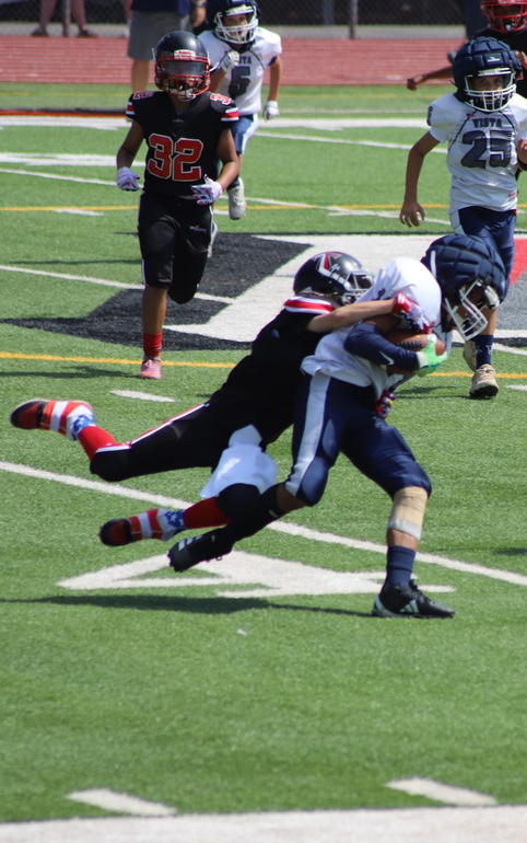 Football players in action on a grassy field; one runs with the ball, pursued by two opponents.