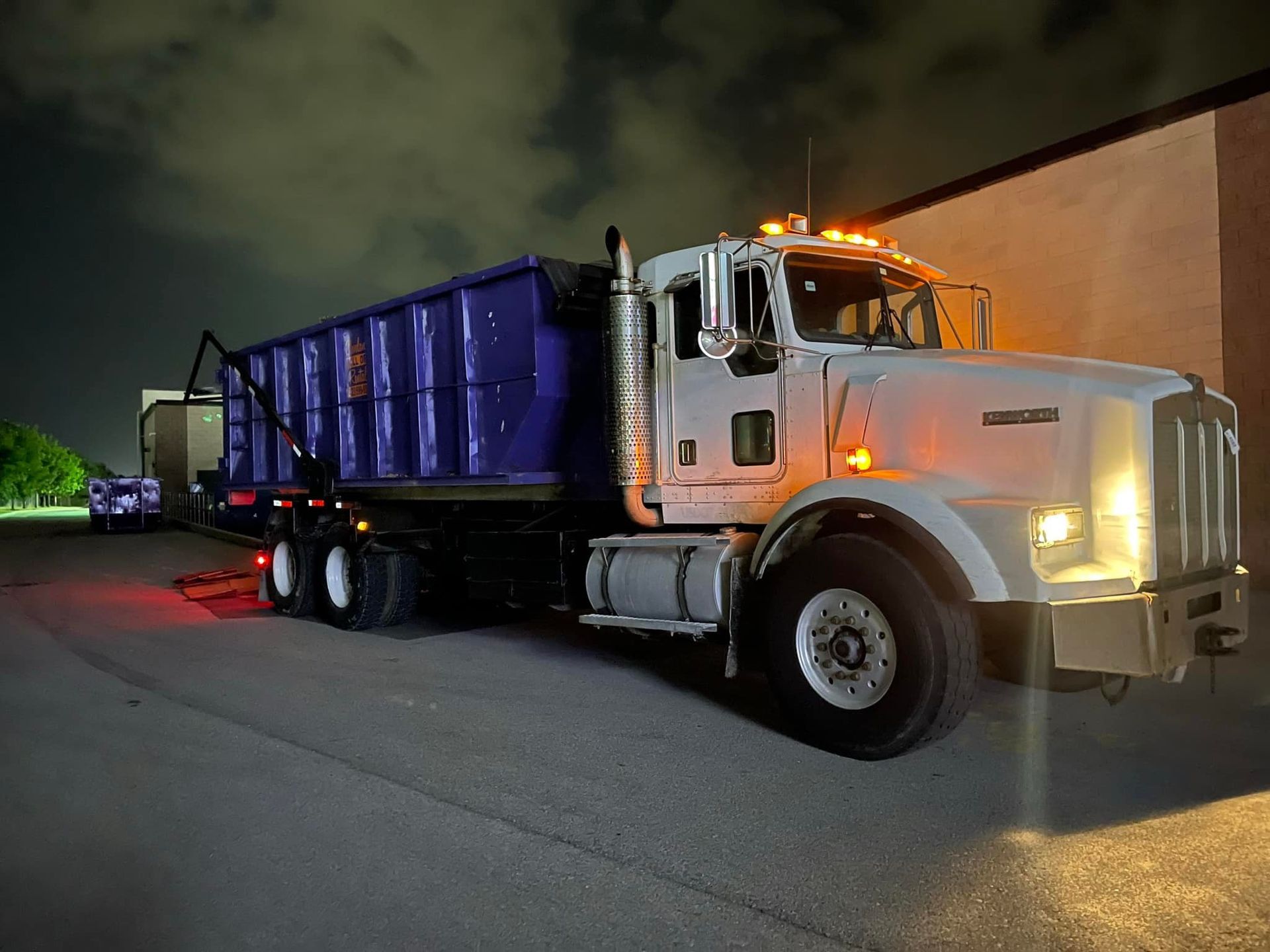 White semi-truck with a blue dumpster parked at night. Headlights and taillights are on.