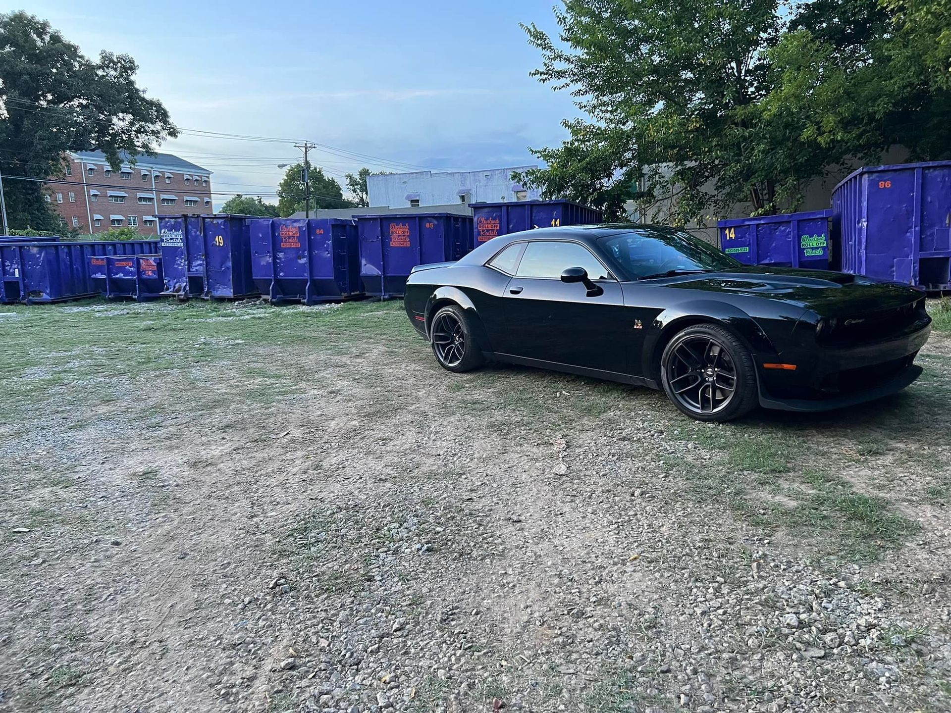 Black Dodge Challenger parked on gravel, with blue dumpsters behind it. Cloudy sky in background.