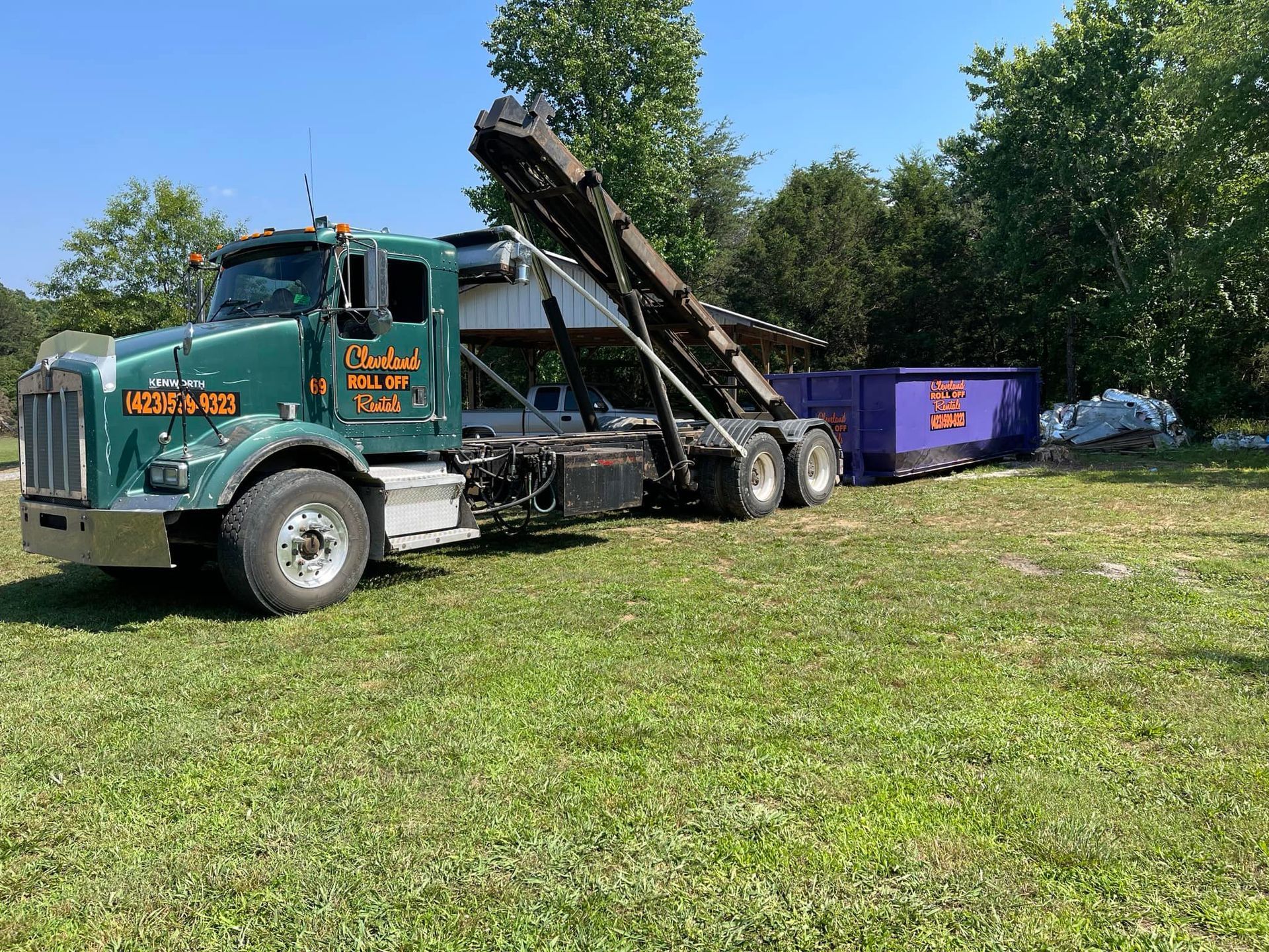 Green and silver truck with a purple dumpster on grass.
