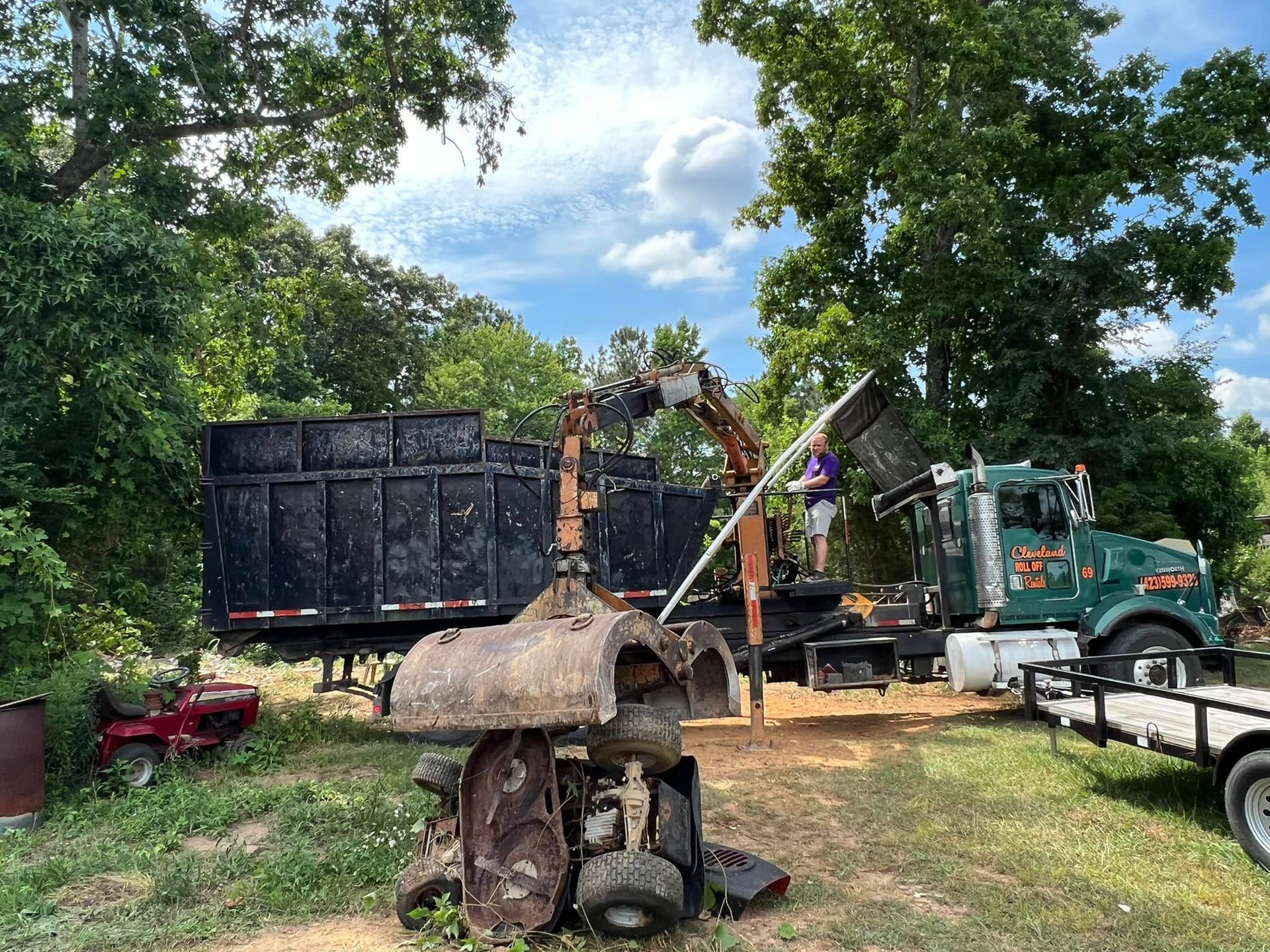 A worker loads debris into a truck bed with a crane. Outdoors, sunny day.