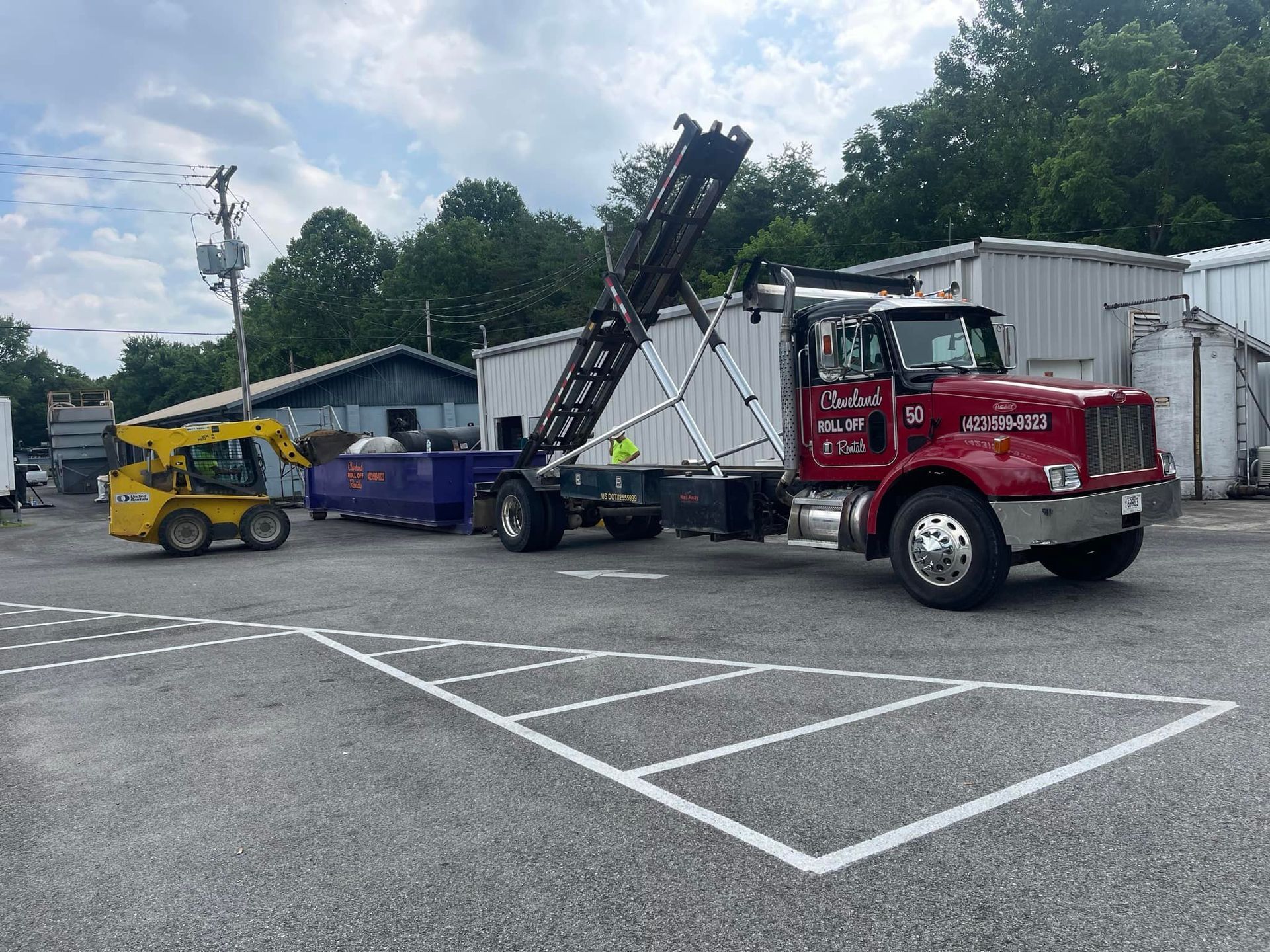 Red dump truck lifting a dumpster, a yellow skid steer in the foreground. Outdoors.