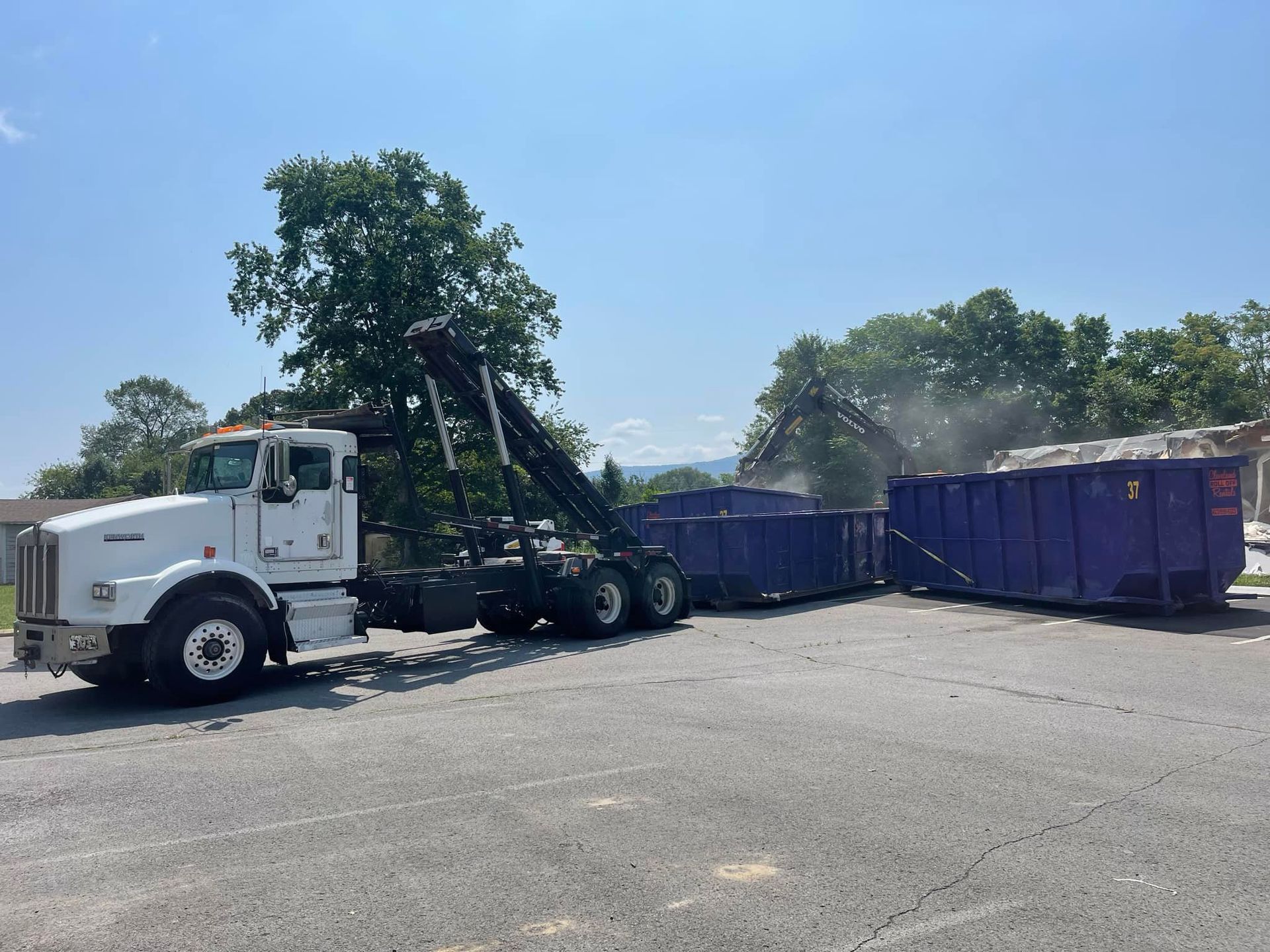 White truck loading two purple dumpsters; construction site.