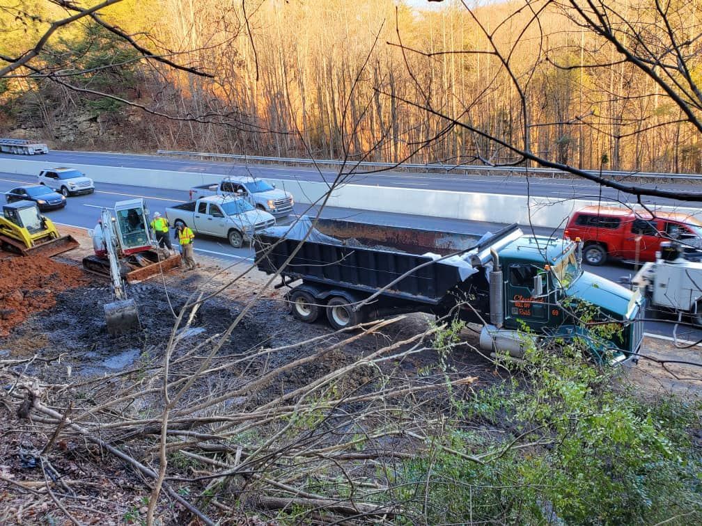Dump truck on side of road, surrounded by workers and vehicles. Construction equipment and cars visible.