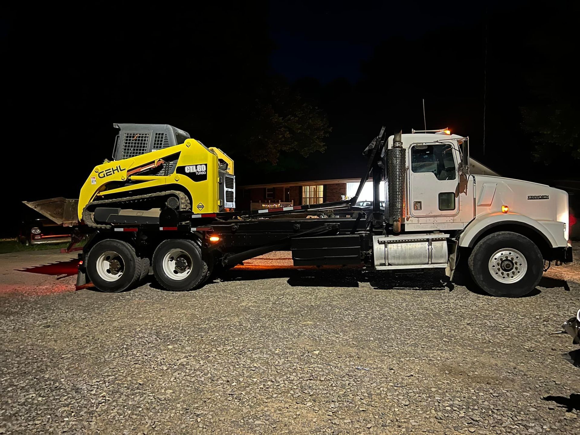 A semi-truck hauling a yellow skid steer loader at night on a gravel surface.