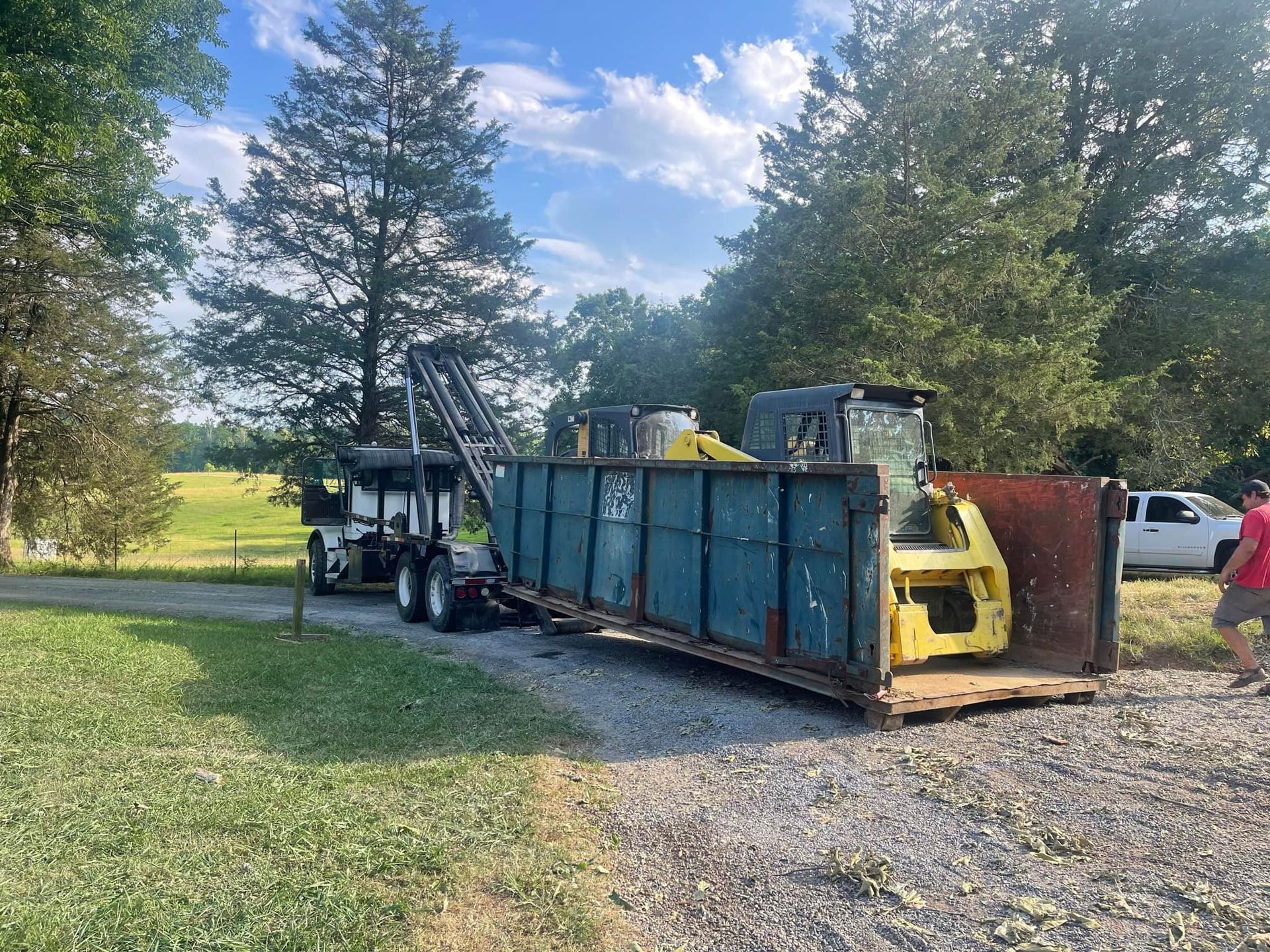 White garbage truck on a road, with a blue dumpster in the back and trees in the background on a sunny day.