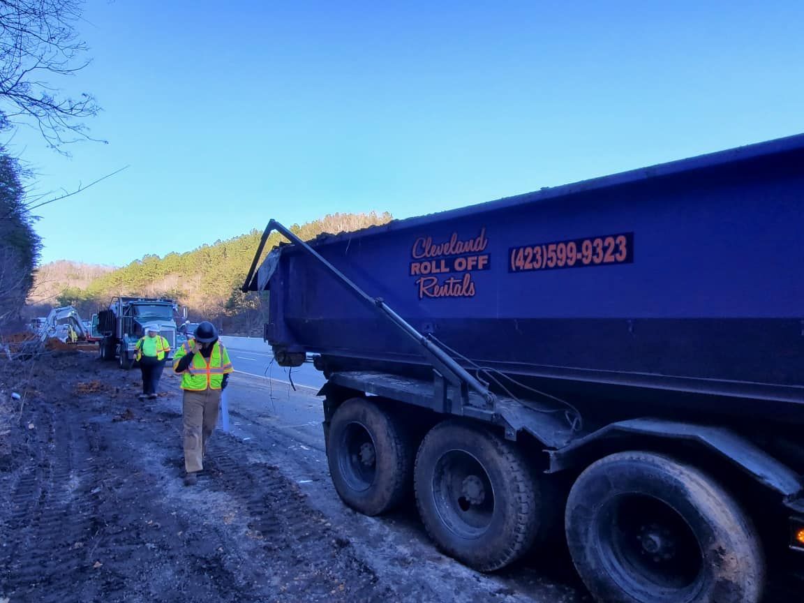 Dump truck on muddy road with people in safety vests. Forest in the background.