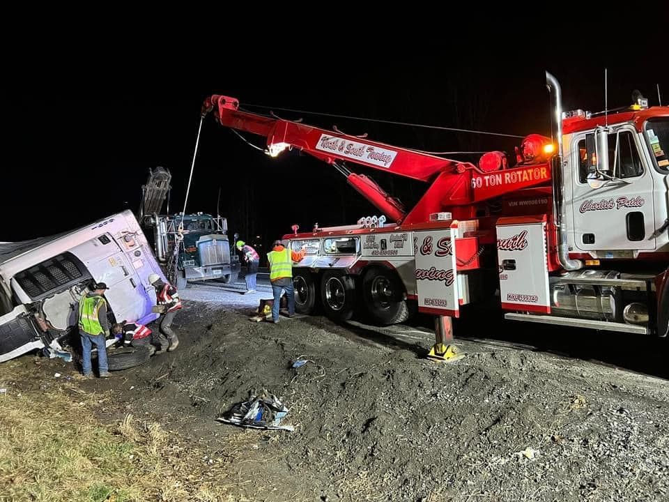 A tow truck at night pulls a bus and truck involved in a crash; emergency responders are present.