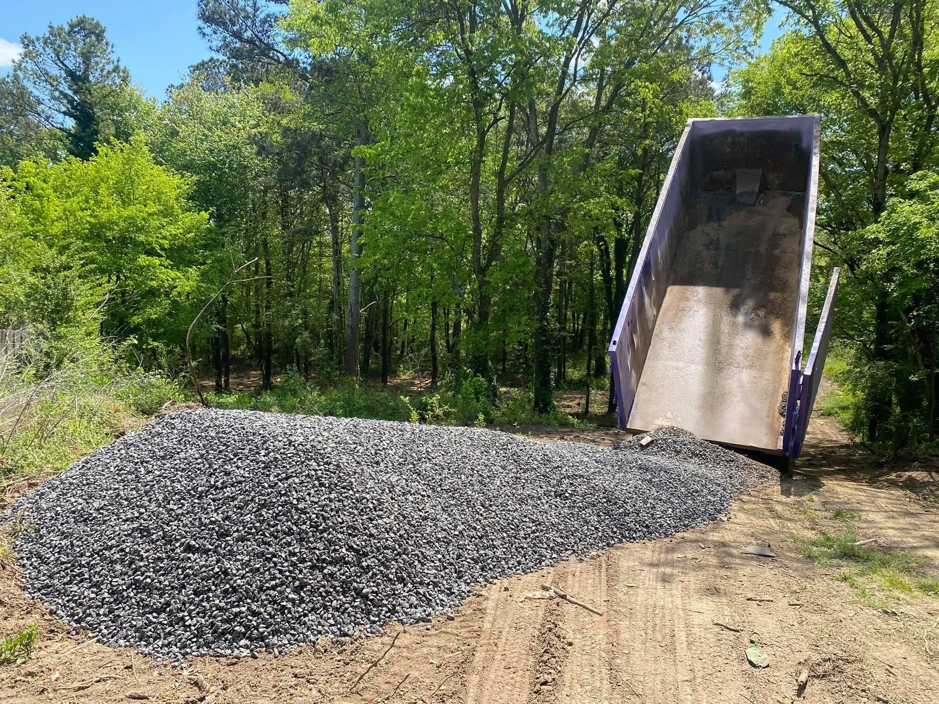 Gravel being dumped from a purple dump truck onto a dirt road with trees in the background.