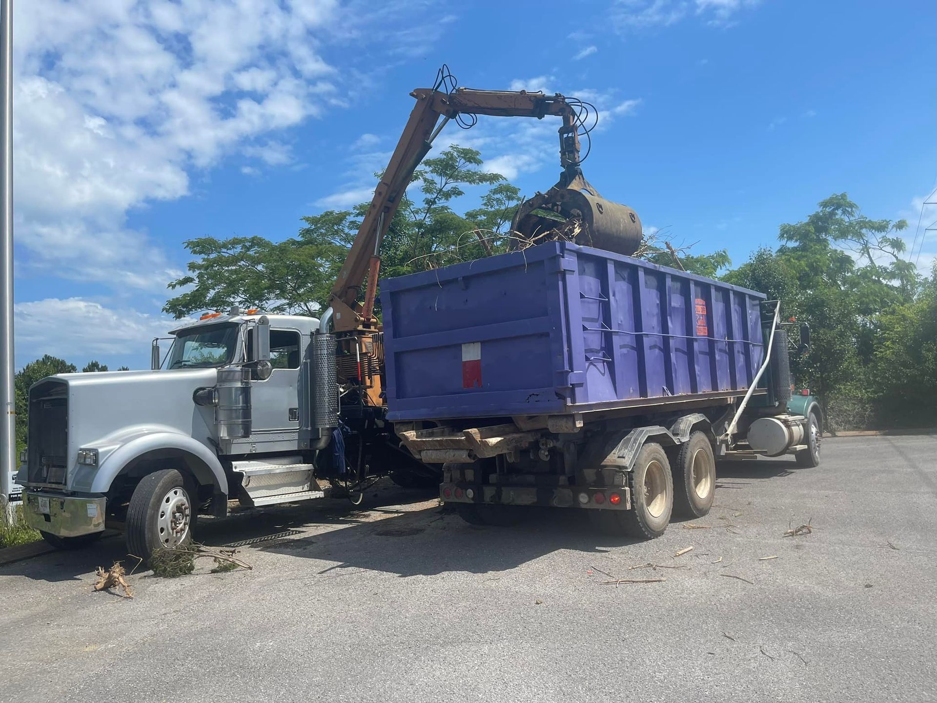 A truck with a crane arm loading debris into a purple dumpster on a sunny day.