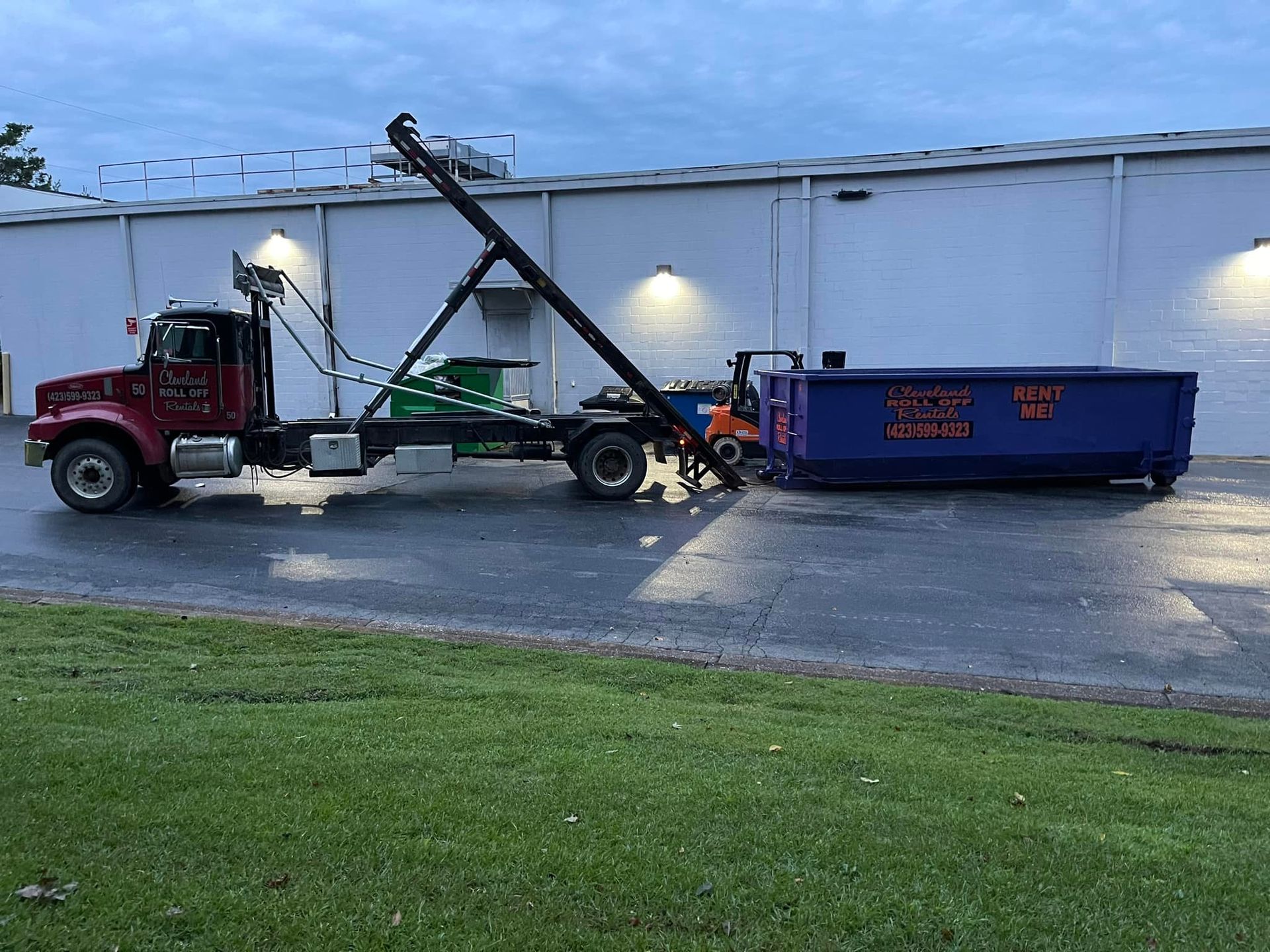 A red truck with a crane lifting a blue dumpster in front of a white building.