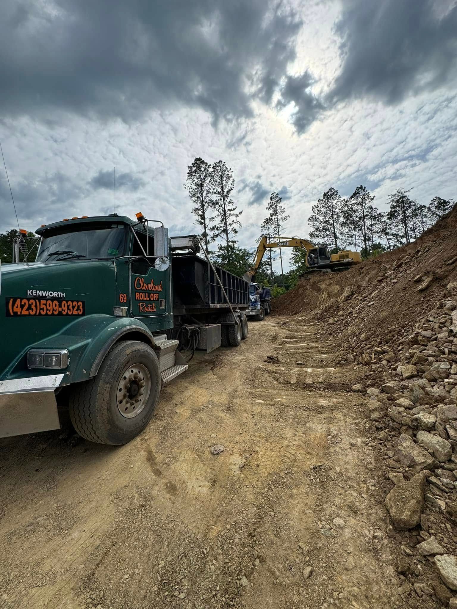Green dump truck parked on a dirt road, excavator in the background, cloudy sky.