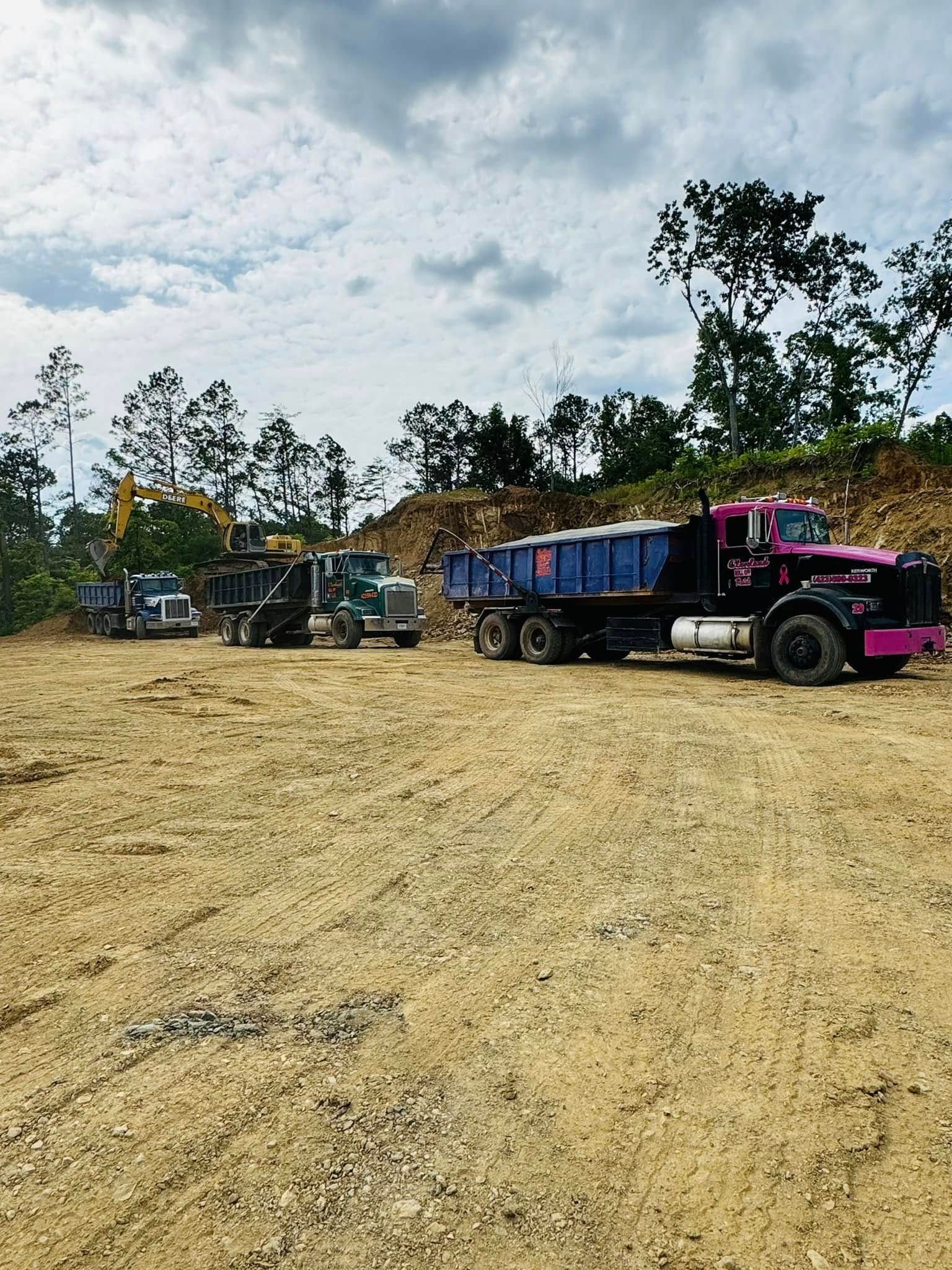 Trucks and an excavator at a worksite; dirt ground with trees and cloudy sky background.