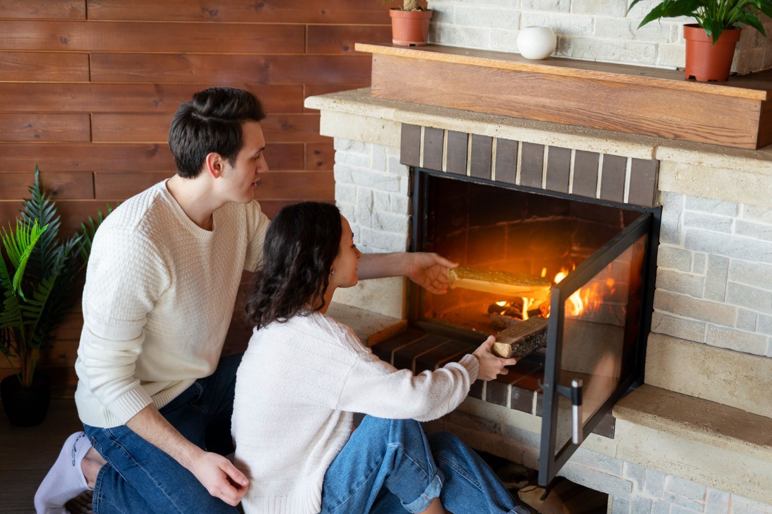 Couple by fireplace, adding wood to the fire. Warm glow, cozy setting, light sweaters.